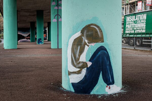 Street art of homeless person in hoodie, seated, at bottom of pillar in Cumberland Piazza, Bristol