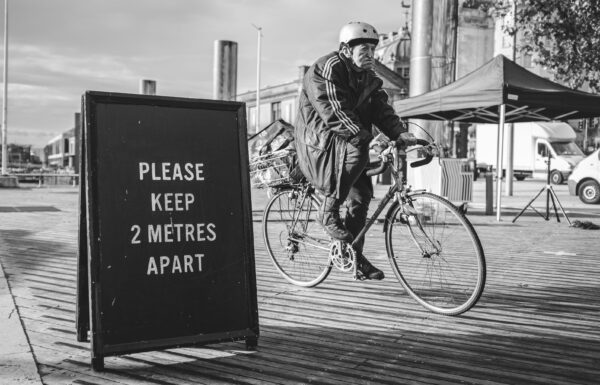 An old man on a bicycle cycles past a PLEASE KEEP 2 METRES APART sign on Bristol's Centre.