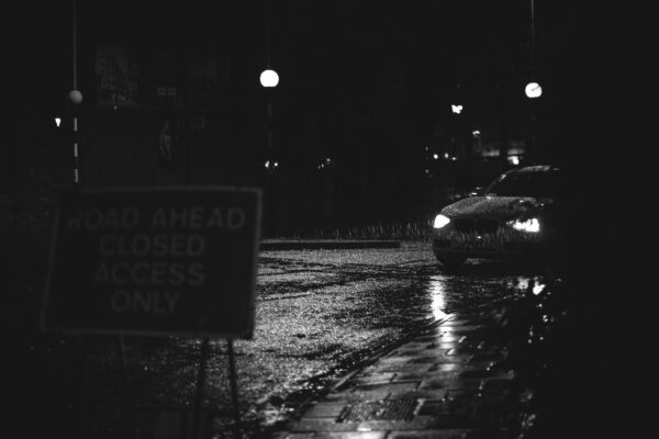 A rainy nighttime scene in Redcliffe, Bristol. A car's headlights pick out raindrops as it approaches a ROAD CLOSED sign.